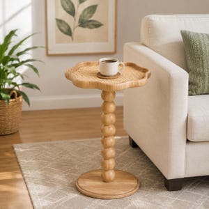 May include: A light brown wooden side table featuring a scalloped, woven rattan tabletop and a central pedestal base composed of stacked wooden spheres. A white ceramic mug filled with dark liquid sits on a small circular coaster on the table.