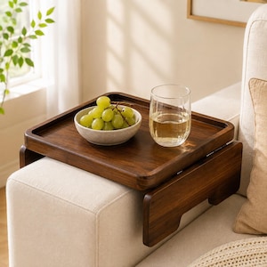 May include: A dark brown wooden sofa arm tray with a rectangular surface and side flaps that drape over the armrest. On the tray sits a small, light-colored speckled bowl filled with green grapes and a clear glass filled with a light-colored liquid.
