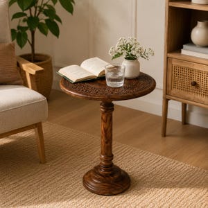 May include: A round, dark brown wooden pedestal side table featuring an intricately carved floral pattern along the edge of the tabletop. The table has a single central column support and a wide, circular base, standing on a textured floor.