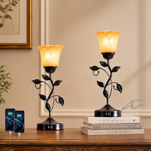 May include: Two table lamps with black metal stems shaped like vines with leaves and amber-colored glass shades. The base of each lamp features a power switch and a USB port. One lamp sits on a stack of three books, while the other stands alone.