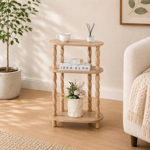 May include: A three-tiered oval side table made of light-toned wood with four spindle-style legs. The top shelf holds a white mug and a glass reed diffuser. The middle shelf features a white book titled IDEA ALPH. The bottom shelf holds a white vase.