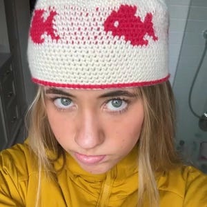 Peut inclure: Un bonnet crocheté de couleur crème orné d'un motif répétitif de petits poissons rouges autour de la calotte. Le bonnet présente une bordure rouge unie le long du bord inférieur, offrant un contraste marqué avec la laine blanc cassé.