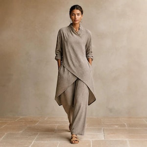 May include: A taupe-coloured long-sleeved tunic with a wrap-style front and asymmetrical hemline, paired with matching wide-leg trousers. The outfit features a textured fabric, a high collar, and simple gold hoop earrings, creating a cohesive look.