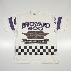 May include: A white short-sleeve t-shirt featuring a Brickyard 400 graphic with the text Indianapolis Motor Speedway, Inaugural Race August 6, 1994. The design includes purple accents, a checkered flag pattern at the bottom, and the number 400.
