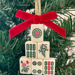 May include: A decorative ornament constructed from six off-white mahjong tiles arranged in a pyramid shape. The tiles feature green, red, and black engraved symbols. A bright red velvet ribbon bow is attached to the top with a gold hanging loop.
