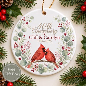 Op de afbeelding: Een rond wit keramieken ornament met een aquarelillustratie van twee rode kardinalen op een tak, omringd door een krans van groene eucalyptusbladeren en rode bessen. De tekst luidt 40th Anniversary, Cliff & Carolyn, 1986-2026.