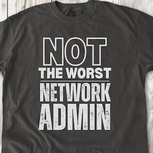 Può includere: Una t-shirt a maniche corte grigio antracite scuro con il testo bianco NOT THE WORST NETWORK ADMIN stampato in un audace carattere sans-serif. La parola ADMIN presenta una texture usurata e invecchiata, mentre le altre righe sono in bianco pieno.