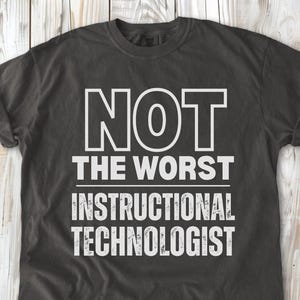 May include: A charcoal gray short-sleeve t-shirt featuring white text that reads NOT THE WORST INSTRUCTIONAL TECHNOLOGIST. The shirt has a crew neckline and a standard fit, with the graphic centered on the chest in a bold, sans-serif font.