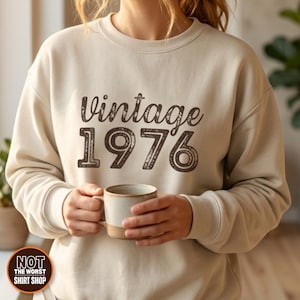 May include: A beige crewneck sweatshirt featuring the dark brown text Vintage 1976 printed on the chest in a distressed font. The sweatshirt has long sleeves with ribbed cuffs and a ribbed neckline, providing a simple and clean appearance.