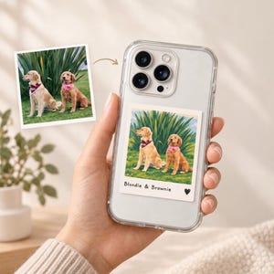 Könnte beinhalten: Eine transparente Smartphone-Hülle mit einem quadratischen Polaroid-Druck von zwei Golden Retrievern, die im Gras sitzen. Der Druck enthält den Text "Blondie & Brownie" sowie ein kleines schwarzes Herz-Symbol am unteren Rand.