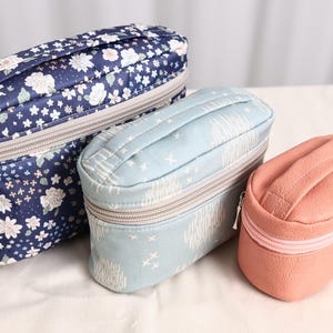 Puede incluir: Tres estuches con cremallera y asa superior. El más grande es azul marino con un estampado floral blanco y azul claro. El estuche mediano es azul claro con un patrón abstracto blanco. El más pequeño es de color rosa salmón liso y texturizado.