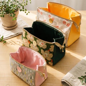 May include: Three open fabric pouches with gold-tone zippers and circular brand tags. The front pouch is gray with a pink interior and colorful triangle patterns. The middle pouch is dark green with floral prints. The back pouch is yellow with a floral pattern.