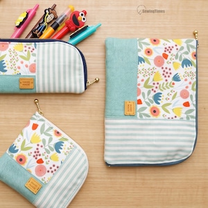 Könnte beinhalten: Drei Stofftaschen mit einem Patchwork-Design aus Blumenmustern, hellgrünen Streifen und einfarbigen türkisfarbenen Abschnitten. Jede Tasche hat ein kleines braunes Lederetikett mit der Aufschrift HAND MADE und einen goldenen Reißverschlussanhänger.