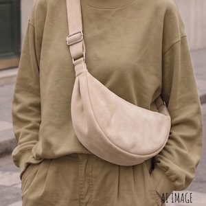 Puede incluir: Un bolso bandolera beige en forma de media luna con textura suave, que cuenta con una correa ancha ajustable y herrajes de metal plateado. El bolso se lleva cruzado sobre el pecho, sobre una sudadera verde oliva de manga larga y pantalones.