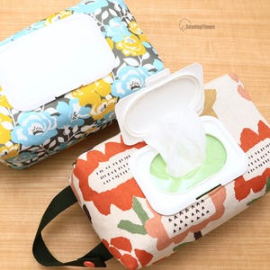 Könnte beinhalten: Zwei rechteckige Feuchttücher-Etuis aus Stoff mit weißen Kunststoffdeckeln. Ein Etui zeigt ein blaues, gelbes und graues Blumenmuster. Das andere hat einen orangefarbenen, grünen und cremefarbenen Blumendruck mit schwarzer Seitenschlaufe.