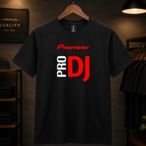 Puede incluir: Una camiseta negra de manga corta con el texto rojo Pioneer sobre el texto blanco PRO y el texto grande rojo DJ centrado en el pecho. La camiseta tiene un cuello redondo y se muestra en una percha de madera sobre un fondo oscuro.