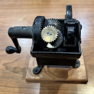 Peut inclure: Un taille-crayon manuel vintage en fonte noire monté sur une base en bois rectangulaire. Le mécanisme comprend un engrenage couleur laiton apparent, une manivelle noire avec un bouton et un système d'engrenages interne complexe.