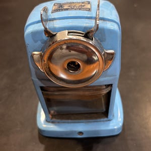 May include: A light blue manual pencil sharpener with a chrome-finished front plate and a clear plastic shavings receptacle. The top features a metal label with the text No 8000, ELM BRAND, and MADE IN JAPAN. The device has two metal wings.