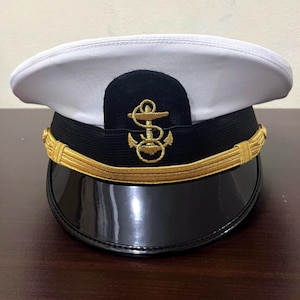Pode incluir: Um quepe branco com uma faixa preta e uma pala preta brilhante. A parte frontal apresenta um emblema de âncora bordado a ouro centrado num remendo preto. Um cordão trançado dourado decorativo atravessa a base da copa, fixado por botões.