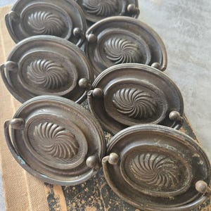 May include: Six antique oval cabinet knobs with a swirl design. The knobs are made of a dark metal with a tarnished finish.