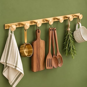 Op de afbeelding: Een wandrek van geborsteld goudkleurig metaal met zeven ronde haken. Aan de haken hangen een gestreepte theedoek, een gouden maatbeker, een houten snijplank, twee houten keukengerei, een bosje verse kruiden en een witte mok.