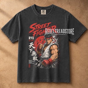 Puede incluir: Una camiseta gris carbón de manga corta con un gráfico del personaje Ryu de Street Fighter en posición de combate. El diseño incluye texto rojo con trazos de pincel, kanji japonés y el texto blanco REBELTREADSTORE en el pecho.