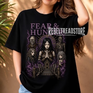Puede incluir: Una camiseta negra de manga corta con un estampado gráfico que incluye el texto FEAR & HUNGER y REBELTREADSTORE. El diseño muestra una figura central con las manos juntas en oración, rodeada por cuatro figuras más pequeñas y humo morado.