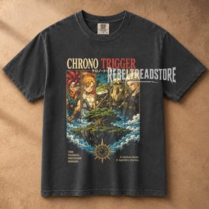 Può includere: Una t-shirt a maniche corte grigio antracite con una stampa grafica dei personaggi di Chrono Trigger, un'isola fluttuante e un emblema a forma di orologio. Il testo recita Chrono Trigger, Time Changes Friendship Remains e A timeless classic.