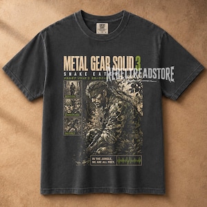 May include: A charcoal gray short-sleeve t-shirt featuring a graphic print of Metal Gear Solid 3: Snake Eater. The design includes a central illustration of a soldier, three smaller square panels labeled Survival, Camouflage, and Instinct, and the text "In the jungle, we are all prey" with a heartbeat line.