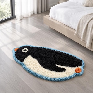 May include: A tufted rug shaped like a penguin, featuring a black body, a white chest, a small orange beak, and an orange foot. The rug is outlined with a light blue border and has a textured, shaggy pile surface throughout the design.