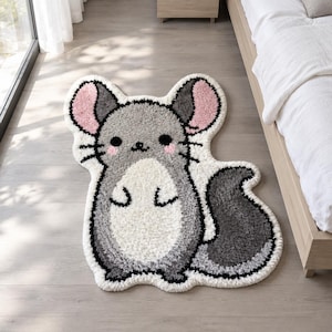 May include: A tufted rug shaped like a chinchilla with a grey body, white belly, and pink inner ears. The design features black eyes, a small nose, whiskers, and a large, curved grey tail. The rug has a thick, textured pile and white border.