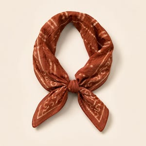 May include: A square scarf in a deep terracotta orange hue, tied in a simple knot. The fabric features a lighter, off-white patterned border and subtle, repeating abstract line designs throughout the surface, creating a textured appearance.