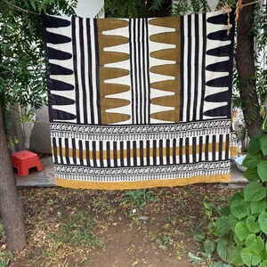 May include: A rectangular textile featuring a geometric pattern in black, white, and mustard yellow. The design includes vertical stripes, repeating pointed shapes, and a horizontal border with intricate floral motifs along the bottom edge.
