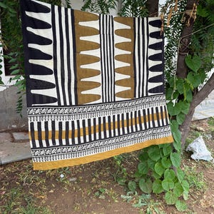 May include: A rectangular textile featuring a geometric pattern in black, white, and mustard yellow. The design includes vertical stripes, repeating triangular shapes, and a horizontal border with intricate leaf-like motifs along the bottom edge.