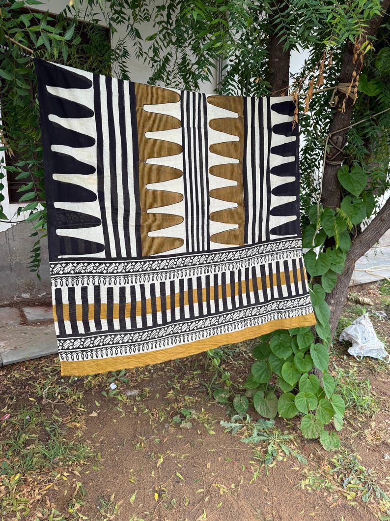May include: A rectangular textile featuring a geometric pattern in black, white, and mustard yellow. The design includes vertical stripes, repeating triangular shapes, and a horizontal border with intricate leaf-like motifs along the bottom edge.