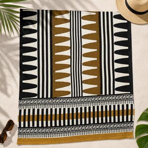 May include: A rectangular beach towel featuring a geometric pattern of black, white, and mustard-brown vertical stripes and repeating triangular shapes. The bottom section displays a horizontal border with intricate leaf-like and striped motifs.