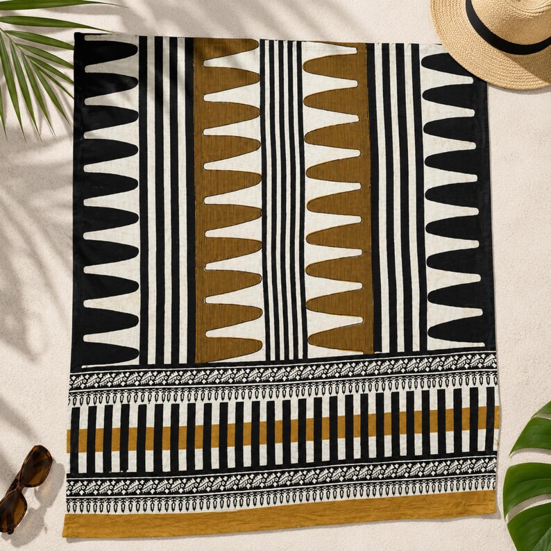 May include: A rectangular beach towel featuring a geometric pattern of black, white, and mustard-brown vertical stripes and repeating triangular shapes. The bottom section displays a horizontal border with intricate leaf-like and striped motifs.