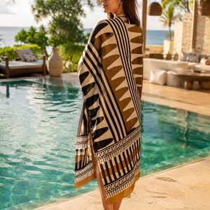 May include: A long, flowing kimono-style cover-up featuring a bold geometric pattern in shades of black, cream, and mustard yellow. The garment displays vertical stripes, triangular motifs, and intricate border detailing along the hemline.
