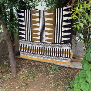 May include: A rectangular textile featuring a bold geometric pattern in black, white, and mustard yellow. The design includes vertical stripes, repeating triangular shapes, and a horizontal border with intricate floral motifs along the bottom edge.