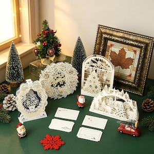 Op de afbeelding: Een verzameling ingewikkelde witte lasergesneden kerstkaarten met vakantiescènes, waaronder een krans, een boom in een raam en een winterlandschap. Inclusief twee kleine kerstmanfiguren, een rood auto-ornament en blanco notitiekaartjes.