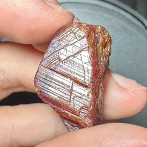 May include: A raw, deep red gemstone with a hexagonal crystal structure and visible striations on its surface. The stone displays a mix of translucent and opaque areas with a natural, unpolished texture, showcasing geometric patterns and facets.