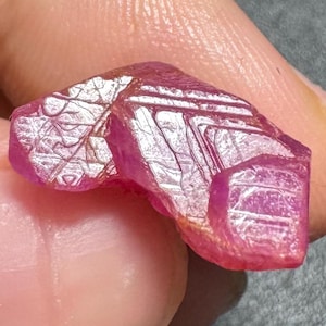 May include: A raw, unpolished ruby gemstone with a vibrant magenta-pink hue. The surface of the stone displays natural, intricate geometric striations and crystalline patterns that catch the light, creating a textured, multifaceted appearance.