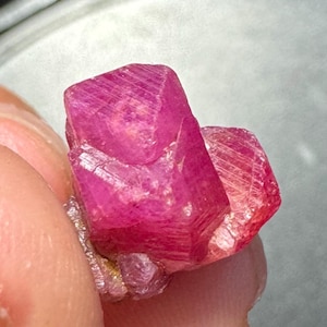 May include: A vibrant, deep pinkish-red raw crystal specimen with a hexagonal, prismatic structure. The mineral features distinct geometric facets, visible striations along its surface, and a semi-translucent quality that catches light.