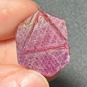 May include: A raw, hexagonal-shaped ruby gemstone with a deep pinkish-red hue. The surface displays a natural, textured crystalline structure with visible geometric facets and internal striations throughout the translucent, vibrant mineral specimen.
