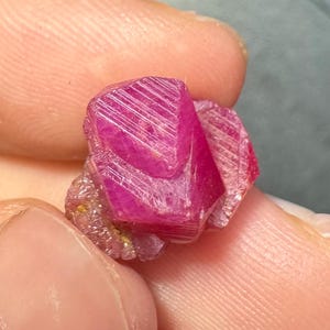 May include: A vibrant, deep pinkish-red raw crystal specimen with a hexagonal, faceted structure. The stone displays distinct linear striations across its surface, with a translucent quality and a cluster of smaller, textured crystalline formations at its base.