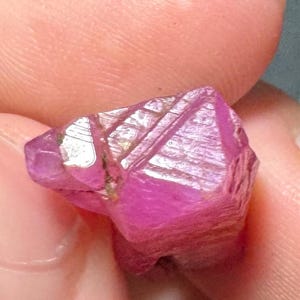 May include: A raw, unpolished gemstone with a vibrant magenta-pink hue. The crystal features a multifaceted, irregular surface with visible natural striations and geometric planes that catch the light, creating bright white reflections across its structure.