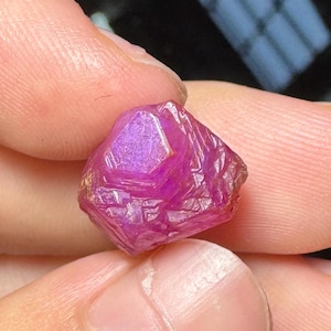 May include: A raw, unpolished gemstone with a vibrant magenta-pink hue. The stone features a natural, multifaceted crystalline structure with visible geometric planes and a translucent quality that catches the light across its textured surface.