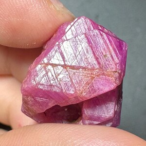 May include: A raw, unpolished ruby crystal with a vibrant magenta-pink hue. The gemstone features a complex, geometric surface with visible striations, natural fractures, and crystalline facets that catch the light, creating a textured appearance.