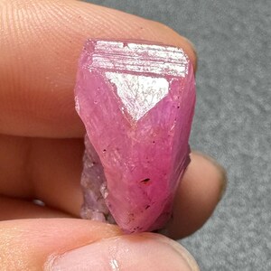 May include: A vibrant pink, translucent crystal specimen with a distinct hexagonal prismatic structure and a flat, polished-looking termination point. The mineral displays natural striations along its vertical faces and a deep magenta hue.