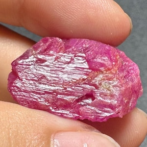 May include: A raw, unpolished ruby gemstone with a vibrant magenta-pink hue. The stone features a rough, crystalline texture with visible striations and natural facets across its surface, displaying varying depths of pink and red coloration.